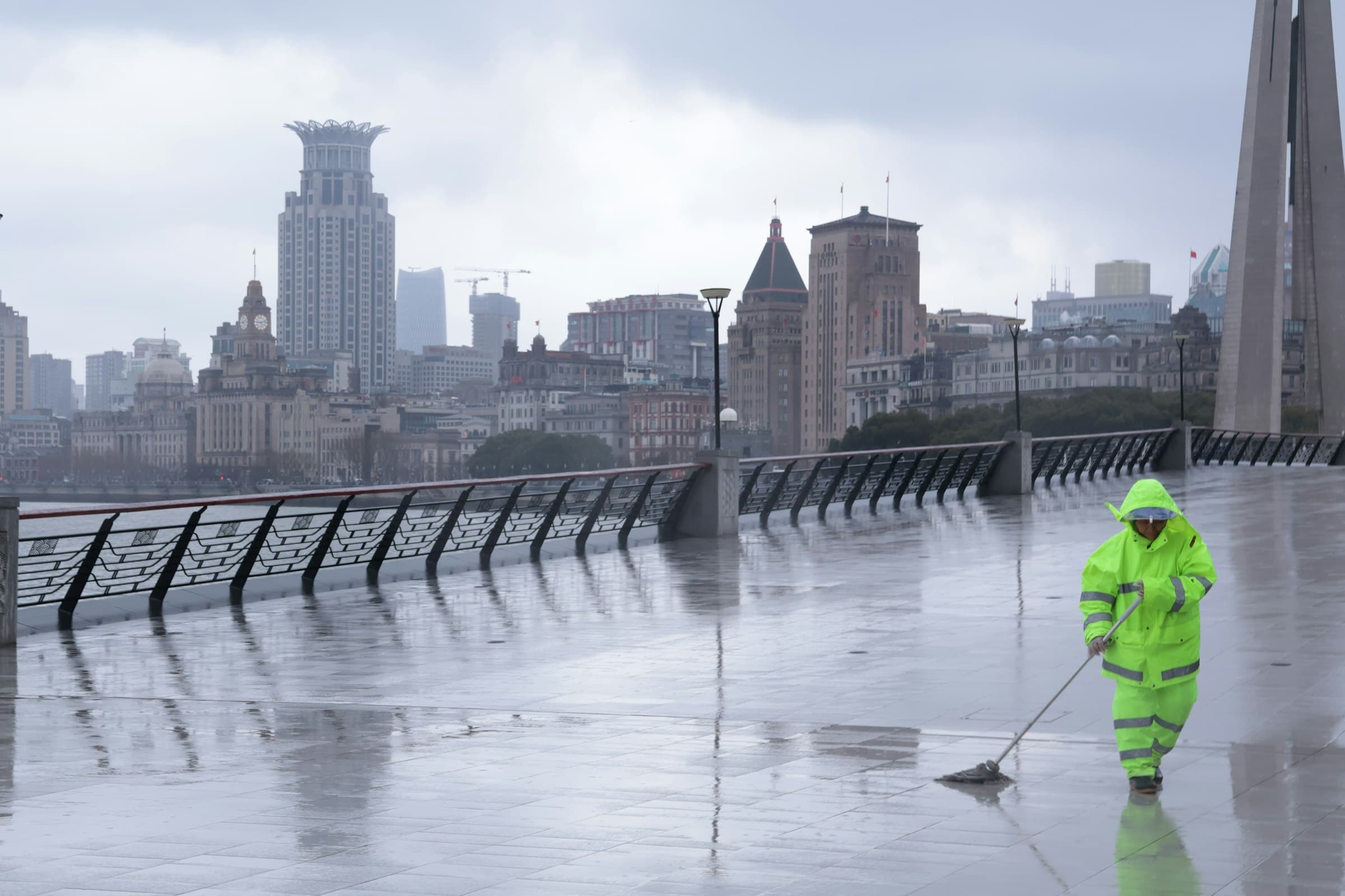 A street cleaner in a bright yellow raincoat sweeps a rain-soaked promenade along the Bund, with Shanghai's historic skyline behind them